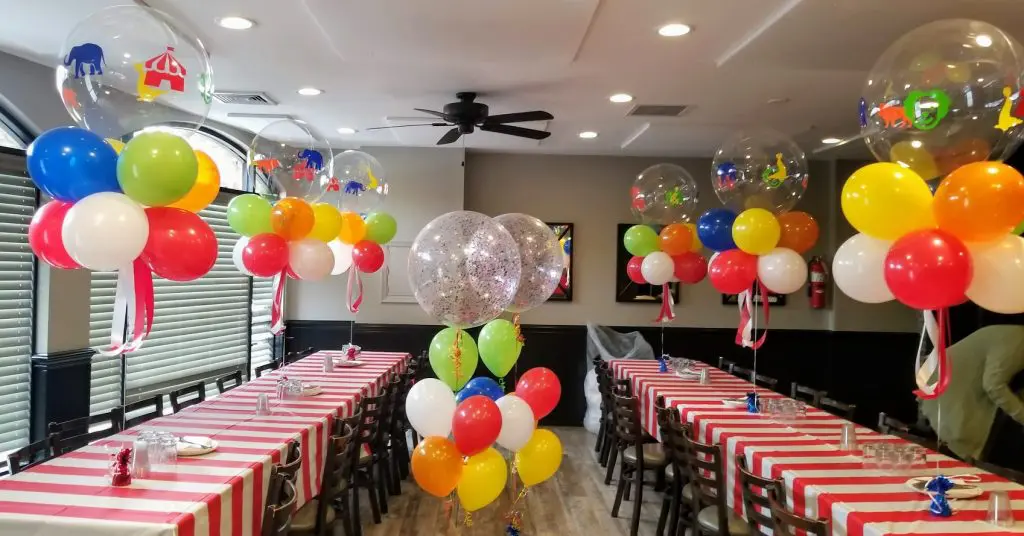 A festive and playful decoration featuring circus theme party balloons in red, green, blue, and yellow colors with confetti accents. The balloons are arranged in a decorative manner and are decorated with circus-themed designs, creating a fun and joyful atmosphere for the event.