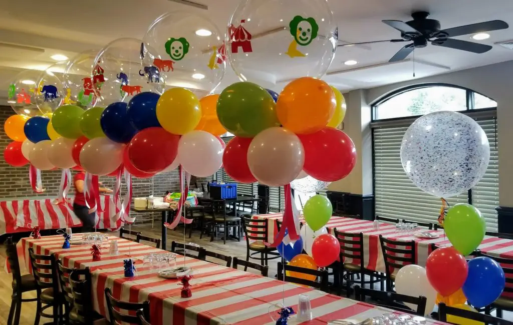 Clear personalized circus theme party balloon table centerpieces, created by Balloons Lane in Brooklyn. The centerpieces feature clear balloons filled with colorful confetti and decorated with circus-themed designs, creating a fun and festive atmosphere for the event.