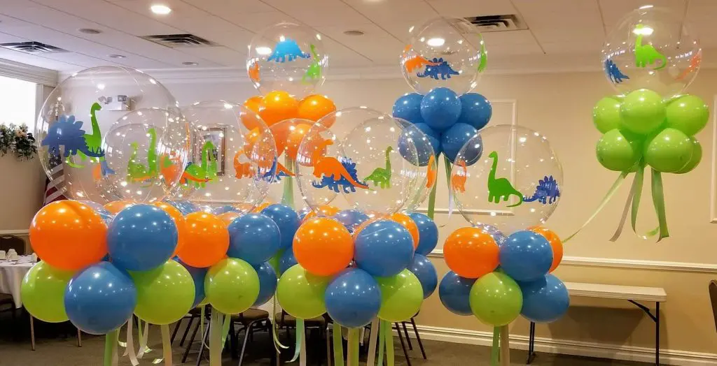 A cluster of clear round balloons with dinosaur-themed prints in shades of green, blue, and orange, arranged as a centerpiece for a 1st birthday party.