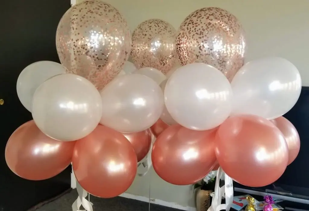 A beautiful and elegant centerpiece for a girl's christening celebration, featuring rose gold and white balloons arranged in a decorative manner. The centerpiece includes confetti balloons, creating a festive and celebratory atmosphere for the event.