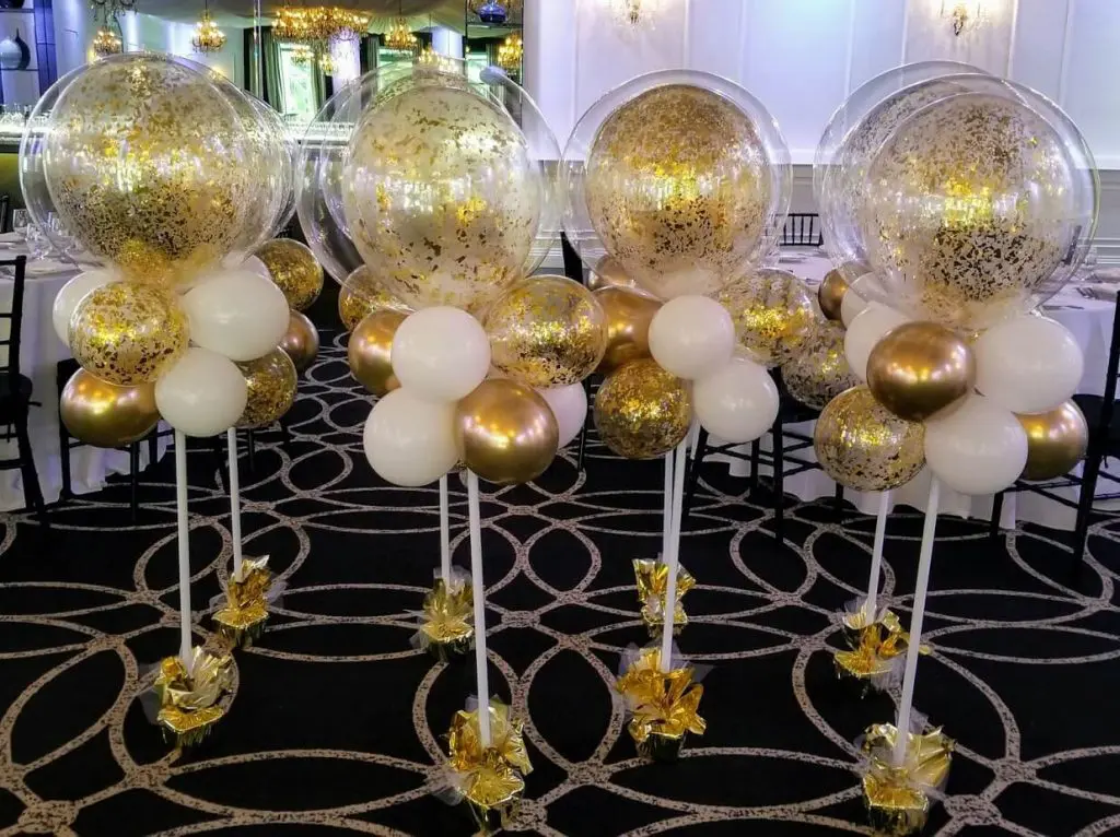 A stunning balloon centerpiece in white and gold confetti balloons, delivered to a location in New York City for an anniversary party. The centerpiece features large, round balloons in the center, surrounded by smaller balloons in a mix of white and gold. The balloons are tied to a weight and appear to be floating in the air, creating a festive and elegant atmosphere for the celebration.