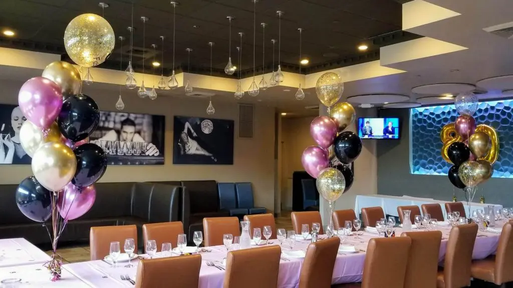 A festive birthday party decoration featuring a metallic gold confetti balloon centerpiece surrounded by a mix of chrome gold, chrome pink, and black latex balloons. The balloons are arranged in a playful and lively manner