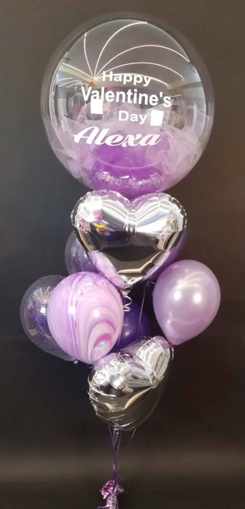 A colorful balloon bouquet in shades of purple, silver, pink, white, and lavender, featuring a large heart-shaped balloon and clear personalized Valentine's Day decorations, in Brooklyn.